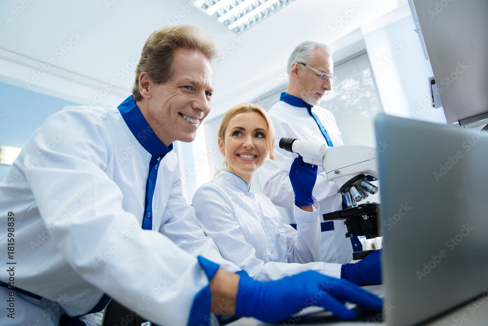 Biologists. Inspired scientist of middle age working on the laptop and a beautiful young woman working with a microscope and smiling while looking at him Biologists. Inspired scientist of middle age working on the laptop and a beautiful young woman working with a microscope and smiling while looking at him
