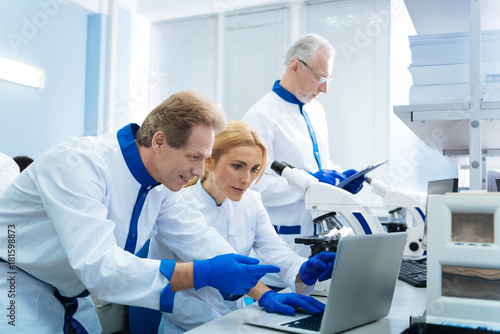 Biology lovers. Smiling alive scientist of middle age working on the laptop and a beautiful blond young woman looking at the screen and an old grey-haired biologist standing in the background