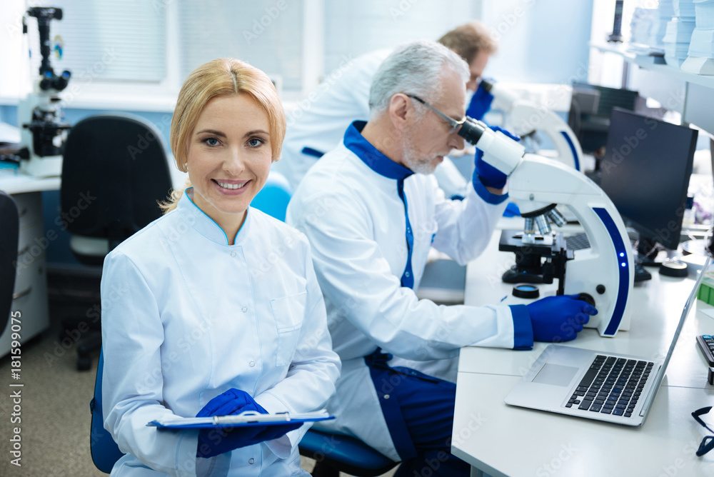 We love genetics. Attractive content blond young researcher smiling and wearing a uniform while other serious researchers working with a microscopes in the background We love genetics. Attractive content blond young researcher smiling and wearing a uniform while other serious researchers working with a microscopes in the background