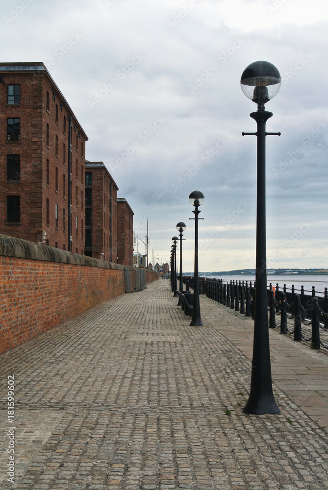 Cobbled paved footpath walkway promenade enbankment along old red brick historic buildings at Liverpool Albert Dock.