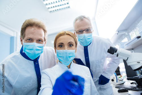 Hey you. Alert vigorous experienced researchers wearing a uniform and a medical face mask and gloves and glasses and the female scientist sitting and men standing near her