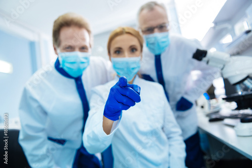 You there. Cheerful vigorous experienced researchers wearing a uniform and a medical face mask and gloves and glasses and the female scientist sitting