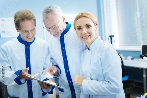 Team spirit. Smiling experienced scientists looking at the screen and wearing uniforms while content female blond researcher standing near them smiling