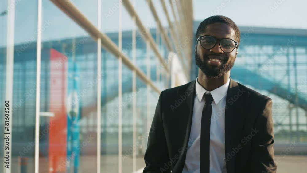 Portrait shot of young attractive African American businessman in a suit and tie posing to the camera and smiling on the big glass modern office center background. Successful man in the city. Outdoor Portrait shot of young attractive African American businessman in a suit and tie posing to the camera and smiling on the big glass modern office center background. Successful man in the city. Outdoor
