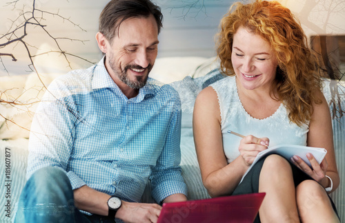 Interesting idea. Positive cheerful friendly couple sitting on the floor next to the comfortable bed and smiling while looking at the screen of a laptop © zinkevych