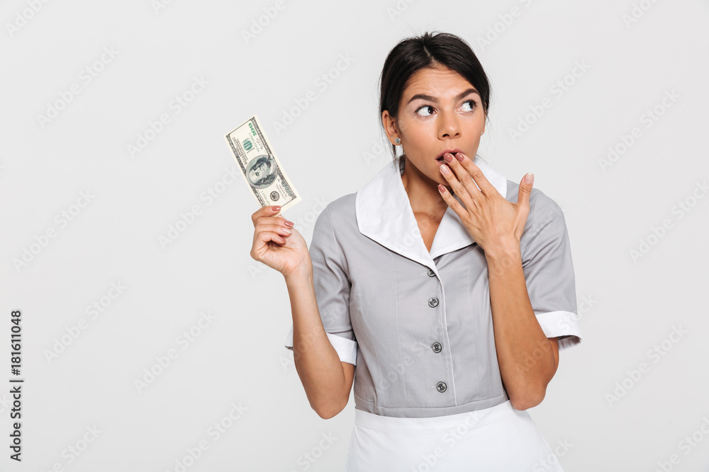 Portrait of amazed attractive maid in uniform holding hundred dollars while covering her mouth with hand, looking aside Portrait of amazed attractive maid in uniform holding hundred dollars while covering her mouth with hand, looking aside