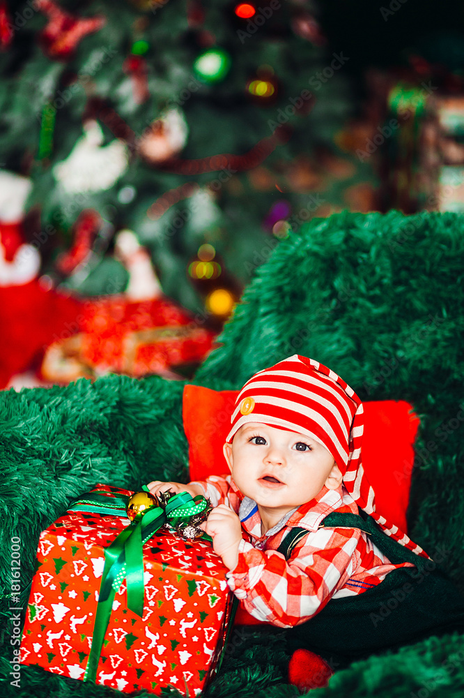 Charming little child in red plaid shirt and Christmas hat sits on green armchair with a present box