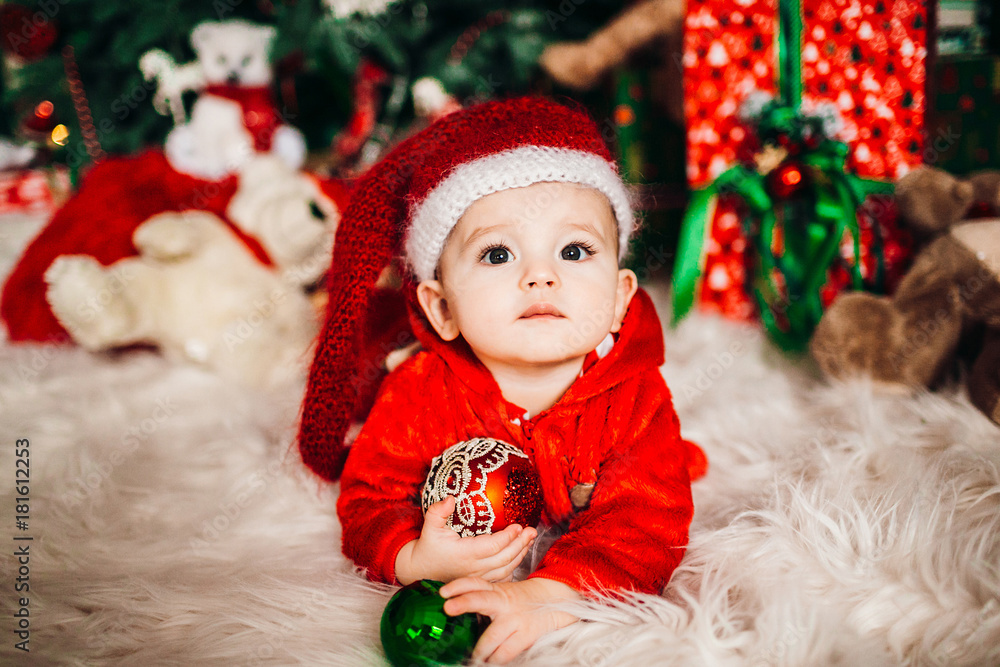 Pretty little boy in red suit lies on fluffy carpet before a Christmas tree Pretty little boy in red suit lies on fluffy carpet before a Christmas tree