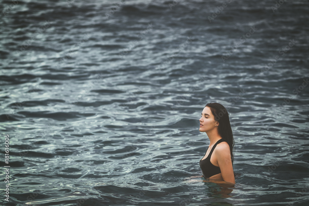 Fototapeta premium Young beautiful girl in dark blue ocean. She is dressed in black swimwear. hipster style, soft light. Empty space for advertising