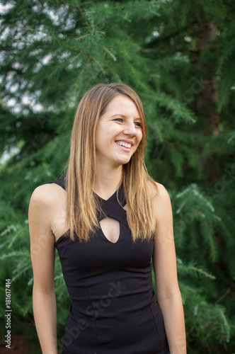 Outdoors Portrait of Attractive Smiling Woman in Black Shirt near Spruce Tree During Summer Time.