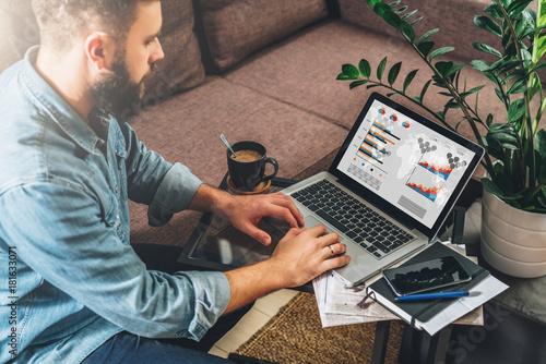 Young bearded hipster man, entrepreneur sits on couch at coffee table,uses laptop with graphs,charts, diagrams on screen.On table notebook,cup of coffee.Online marketing,education,e-learning. Startup.