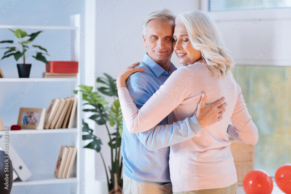 Fototapeta premium Dozens of years together. Joyful husband and wife closing their eyes and smiling while dancing at home and enjoying the moment together.