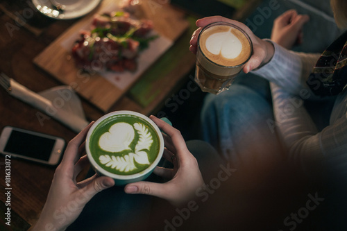 Two friends meet for reunion at local coffee shop or cafe spot to talk and share gossip, enjoy macha latte with milk and hot chocolate, with pretty art drawing by professional barista