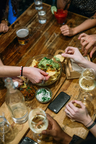 Group of friends share meal of tapas and snacks at big wooden table at bar or restaurant. take out crispy corn tortillas or nachos and dip them in guacamole, celebrate with drinks and cocktails