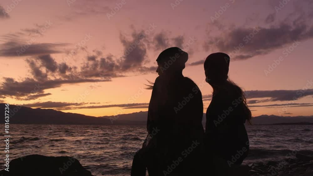 Two young women sitting next to lake enjoying the sunset on cold Autumn evening. Two young women sitting next to lake enjoying the sunset on cold Autumn evening.