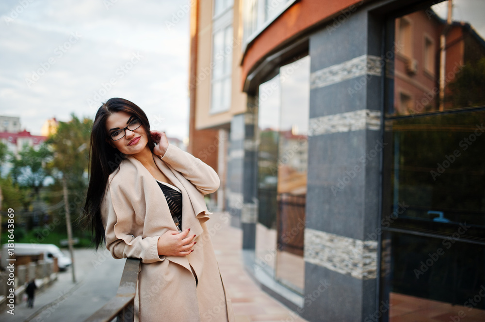 Black hair sexy woman in glasses and coat posed against building with modern windows.