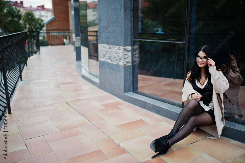 Black hair sexy woman in glasses and coat posed against building with modern windows.