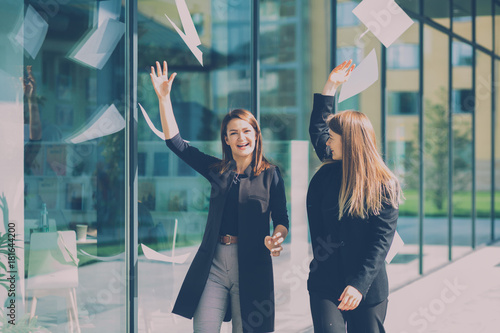 Photos Two happy business woman throwing documents in the air while going along modern building