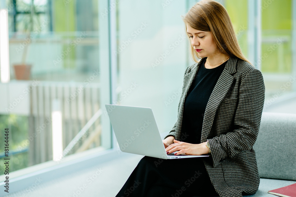 Young business woman sitting at sofa and working with laptop at the waiting hall of the business center. Young business woman sitting at sofa and working with laptop at the waiting hall of the business center.
