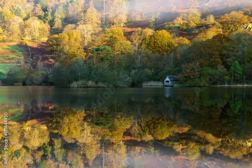 Boathouse at Rydal Water lake with clear calm Autumnal reflections and bright sunlight.