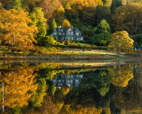 Fototapeta Naklejka Na Ścianę i Meble -  Rural country house reflecting in a calm lake with stunning Autumn colours. Taken at Rydal Water in the English Lake District.