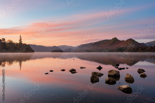 Beautiful pink sunrise reflected in a calm misty lake with rocks in foreground. Taken at Derwentwater in the English Lake District.