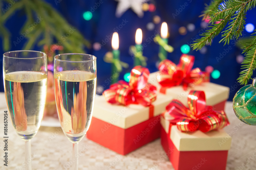 on the table glasses filled with champagne on a dark blue background with blurred lights. near there are New Year's gifts decorated with red bows. a fragment of the New Year tree on the table glasses filled with champagne on a dark blue background with blurred lights. near there are New Year's gifts decorated with red bows. a fragment of the New Year tree