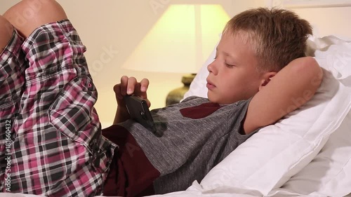 Closeup video of cute blond kid laying on back in bed at home interior. Child holds modern smart phone and playes cheerfully videogame. Real time full hd video footage. photo