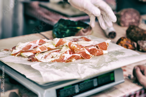 Close-up of thin slices of Italian ham, prosciutto crudo weight on scale, grocery store, hands of seller, vintage. Selective focus