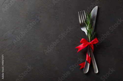 Festive set of cutlery knife and fork with red satin bow with rosemary, dark stone slate background, top view, copyspace, horizontal image
