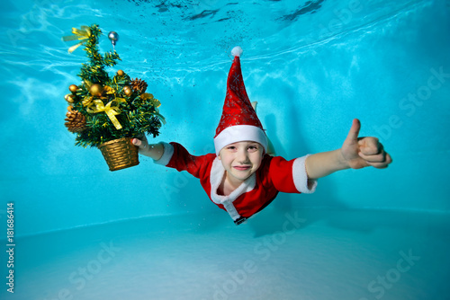 A little boy in a cap Santa Claus swimming underwater with outstretched arms, and with a Christmas tree in hand on blue background, looking at camera and smiling. Shooting under water