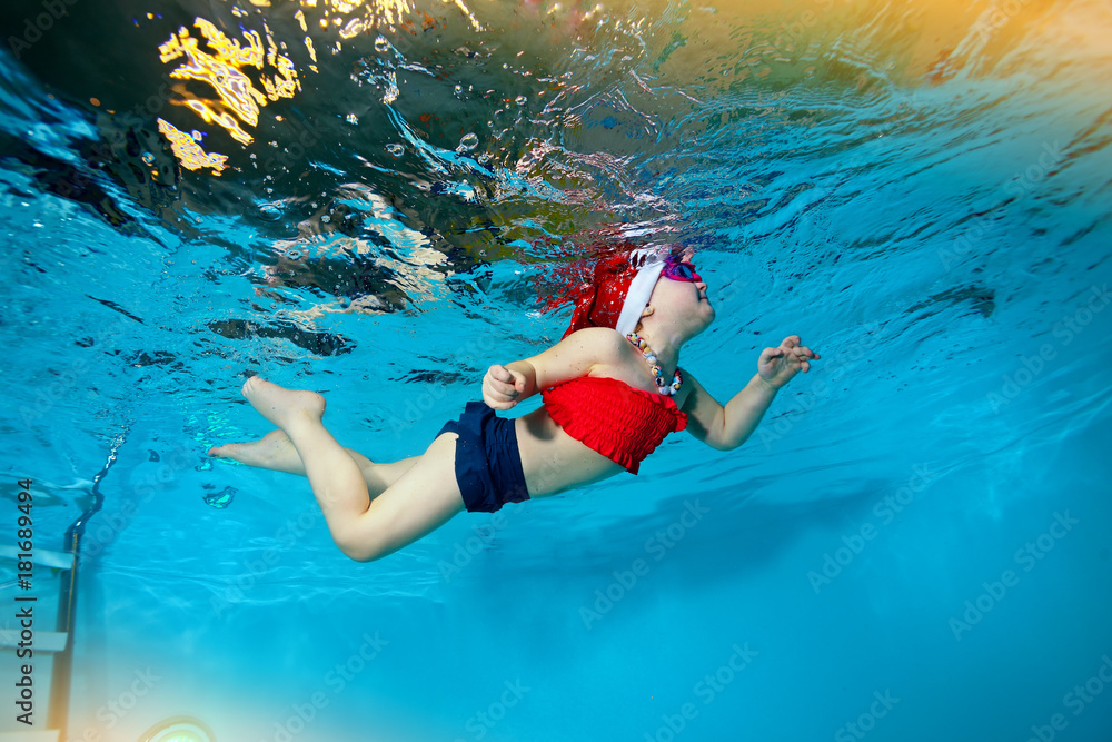 The little girl floats underwater in a cap of Santa Claus in the pool on a blue background. Portrait. Shooting under water. Horizontal orientation