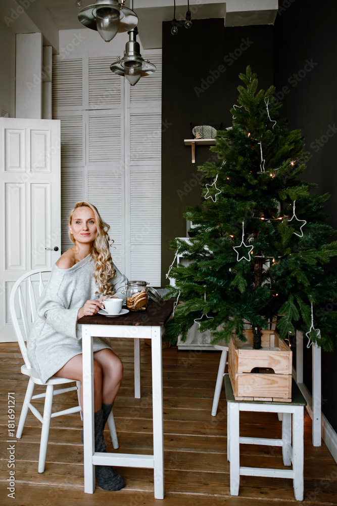 Christmas morning. The beautiful young woman the blonde in a winter white sweater has tea or coffee in kitchen against the background of a Christmas fir-tree.