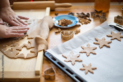 Close up photo of female hands and wooden board with a dough and baking pan with raw cookies in a shape of star. Woman is cutting a christmas cookie with cookie cutter in the shape of star.