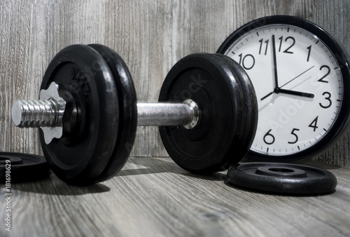 Black dumbbell on wooden background with wall clock.