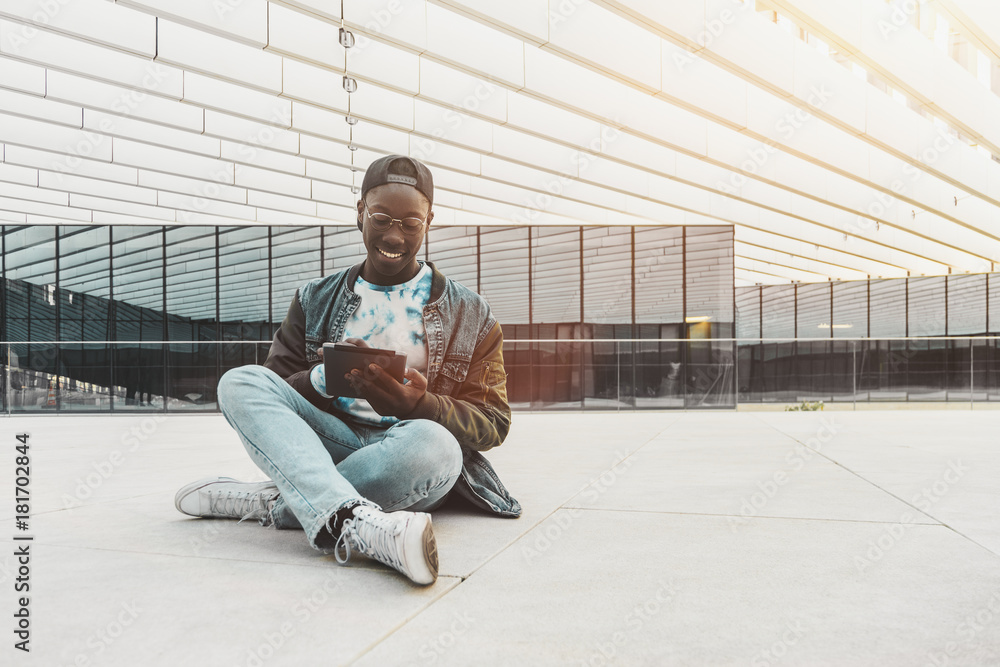 Cheerful young black student is sitting on the stony floor outdoors in modern university campus and having online chat with his friends via tablet pc with copy space place for advertising or your logo Cheerful young black student is sitting on the stony floor outdoors in modern university campus and having online chat with his friends via tablet pc with copy space place for advertising or your logo