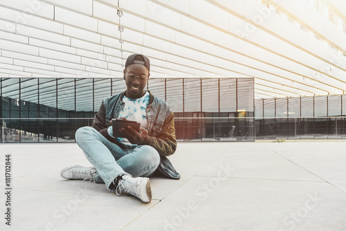 Cheerful young black student is sitting on the stony floor outdoors in modern university campus and having online chat with his friends via tablet pc with copy space place for advertising or your logo