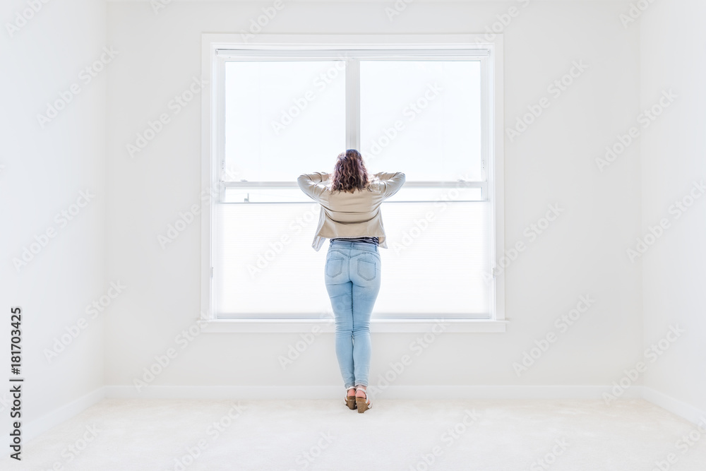 Back of one young woman standing in white, bright empty, clean room on carpet of modern new house, home, apartment looking through the sunny large window with blinds Back of one young woman standing in white, bright empty, clean room on carpet of modern new house, home, apartment looking through the sunny large window with blinds