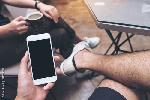 Mockup image of hand holding white mobile phone with blank black screen on thigh with white canvas shoes in cafe