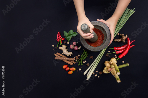 The Art of Thai Cuisine - Thai lady’s hands hold stone granite pestle with mortar and red curry paste ingredient together with fresh herbs and spices on classic dark background at top view angle. photo