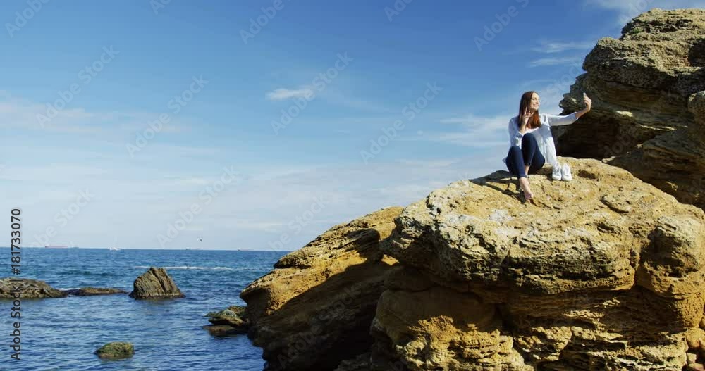 Beautiful young barefoot woman sitting on the big cliff and taking selfies on the smart phone on the cliffed sea background. Chilly sunny day. Outdoors Beautiful young barefoot woman sitting on the big cliff and taking selfies on the smart phone on the cliffed sea background. Chilly sunny day. Outdoors