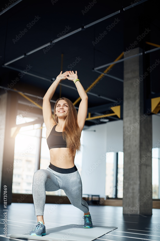 Obraz premium Beautiful female fitness idol making squats smiling early in the morning in empty gym. Portrait of fit woman training hard.