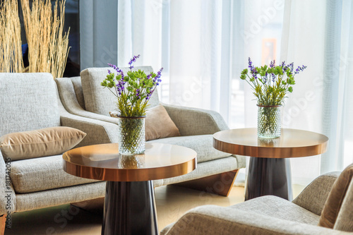Green flowers and purple Flowers bouquet in glass vase on table with fabric sofa and blinds in living room.