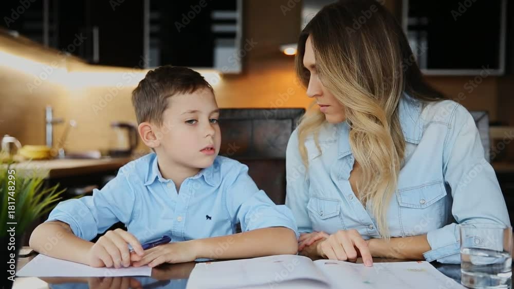 Smiling mother helping his son doing homework, dictates the text to help you write sitting at the table in the kitchen. Smiling mother helping his son doing homework, dictates the text to help you write sitting at the table in the kitchen.