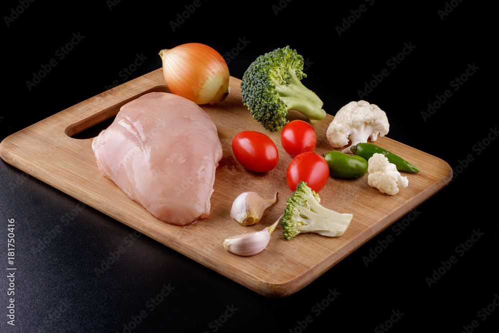 Raw chicken breast fillet with broccoli, green chillies, onion, tomatoes on a dark background.