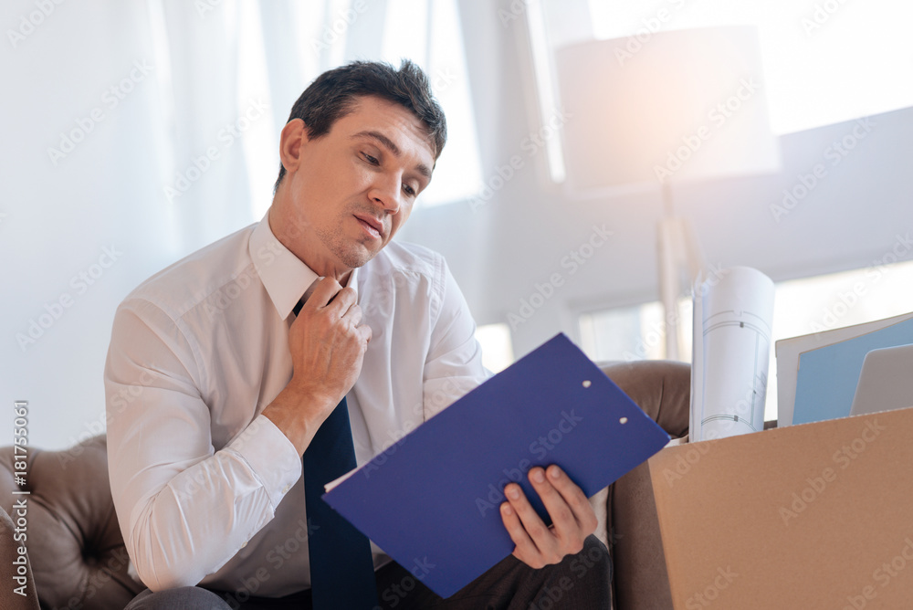 Old documents. Serious angry attentive man looking at the documents in a blue clipboard while sitting in a comfortable armchair after losing his job Old documents. Serious angry attentive man looking at the documents in a blue clipboard while sitting in a comfortable armchair after losing his job