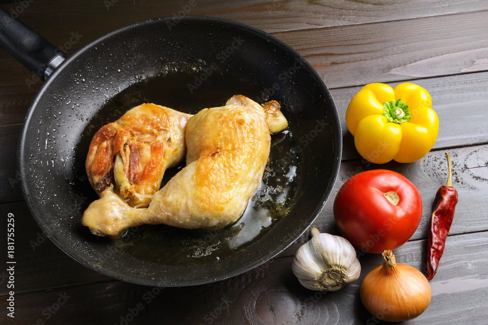 Two roasted chicken legs in black frying pan with vegetables and spices on dark wooden planks Two roasted chicken legs in black frying pan with vegetables and spices on dark wooden planks