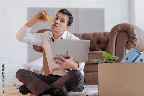 Tasty. Calm attentive young jobless man looking at the screen of his laptop while sitting on the floor with his legs crossed and drinking alcohol from the bottle