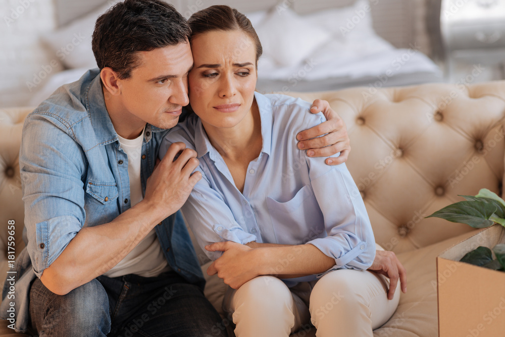 Better. Attentive kind loving man sitting on the sofa and hugging his sad unhappy dismissed wife while trying to encourage her Better. Attentive kind loving man sitting on the sofa and hugging his sad unhappy dismissed wife while trying to encourage her