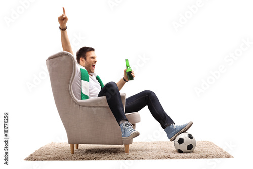 Cuadro en lienzo Excited football fan with a scarf and a beer bottle seated in an armchair pointi